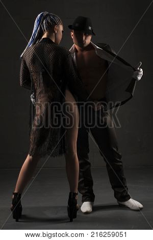 Young girl with African braids and sexy dressing gown staying close to her boyfriend wearing suit and hat view near black wall
