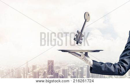 Closeup of waiter's hand in white glove presenting stone key symbol on metal tray with cityscape view on background. 3D rendering.