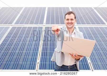 A handsome man of European appearance keeps an open modern laptop in his hands, smiling and showing his index finger in front of himself, standing on a background of solar panels. Outdoors