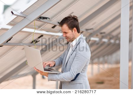 A smiling man of European appearance checks the work of solar panels through a modern program on a laptop. Outdoors