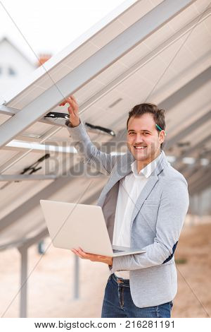 A young man holds an open modern laptop in his hands, and holds his hand behind the solar panel and smiles. Outdoors .