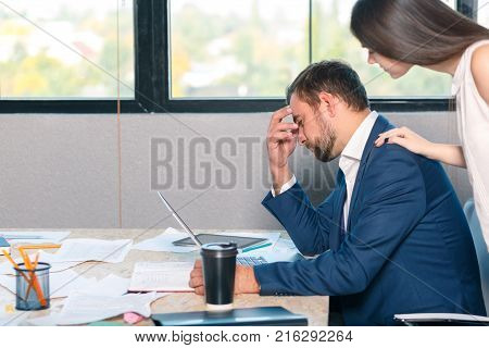 A sad man, an office worker, is sitting up at the desk frantically and holding his head, a young employee stands beside him and comforts and supports him. Inside the office.
