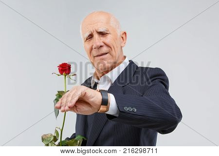 A handsome mature man in a black suit, looks at his wristwatch and durts in his hand one red rose. Isolation.
