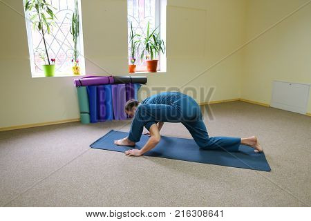 Beautiful dark-haired man performs asanas from yoga, guy sitting in room on floor on fitness mat. Inside spacious and bright, walls painted in nice yellow color, on windowsills pots with flowers. Concept of importance of keeping body in good shape, exerci