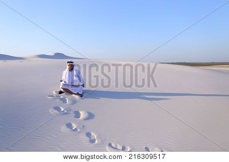 Beautiful Emirate Sheikh businessman studying project of future construction of complex, standing in middle of bottomless desert with white clean sand on clear warm day against blue sky. Swarthy Muslim with short dark hair dressed in kandura, long, spacio
