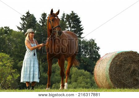 Woman in blue dress stay beside her horse near hayroll
