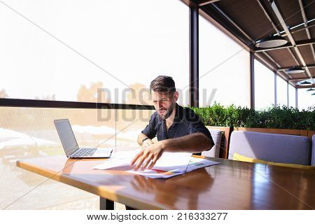 Placeman sorting papers on desk near laptop. Strong male hands with watch near window. Concept of stopping document clutter on workplace.