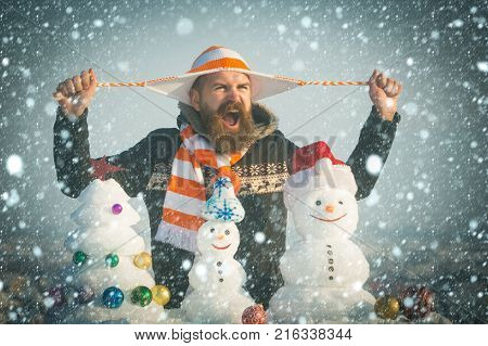 new year christmas snow concept Angry man holding hat strings in hands. Holidays celebration concept. Hipster shouting on winter day. Christmas and new year. Snowmen and snow xmas tree on blue sky.