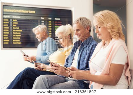 Elderly people are sitting in the waiting room at the airport. They look at their smartphones. There is a timetable on a background.