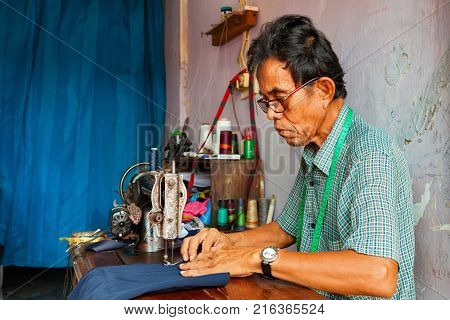Jakarta Java island Indonesia - August 29 2015: A way of life earning money in Jakarta city. Portrait of senior tailor man at work - sewing clothes on old sewing machine at street tailoring stall.
