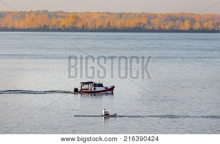 Early morning rowing exercise with a red boat passing on Lac St Louis, Lachine, Quebec Canada, in late fall with the golden trees illuminated on the far side of the lake.
