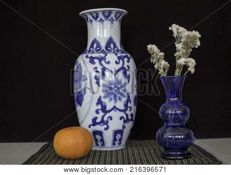 Blue glass vase and oriental vase still life on striped cloth set against dark background.