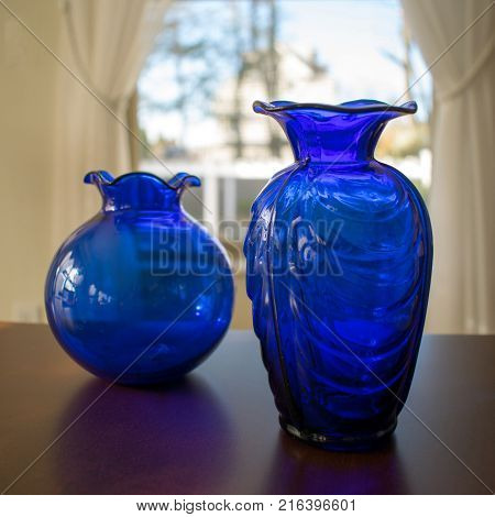Two shiny blue vases in front of a window with reflections on wood table.