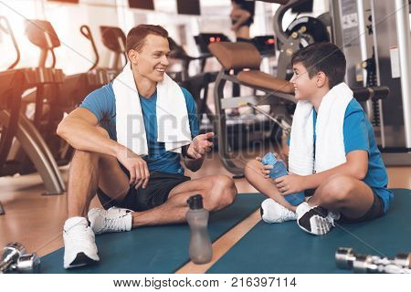 Father with his son in the same clothes in the gym. Father and son spend time together and lead a healthy lifestyle. The man and boy took a break during training. They have bath towels around necks.