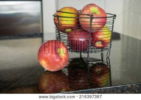 One apple beside a metal basket filled with red apples and strong reflections.