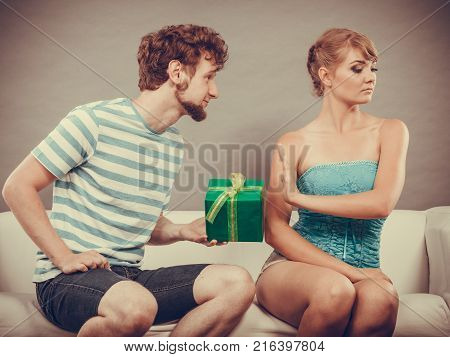 Couple sitting on couch at home. Young man giving offended woman gift box girl refusing to accept present