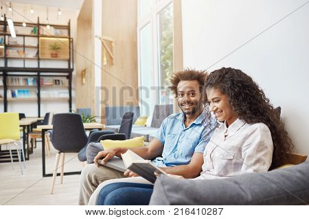 Close up of two young people in casual clothes sitting on sofa in modern library. Girl reading to book, getting ready for universit lecture. Guy looking in camera with happy face expression, holding book in hands