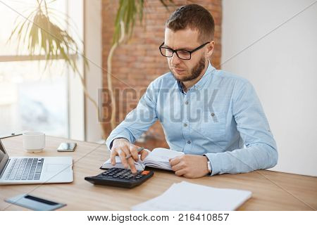 Adult serious bearded caucasian finance manager in glasses and blue shirt sitting in light comfortable company office, cheking month profits with calculator, writing down results in notebook, speding productive morning