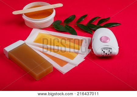 Close-up of a female epilator, wax strips, wax, wax in a cassette, lie on a red background, next to it there is a green twig.