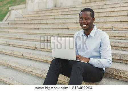 Happy black businessman working on laptop. African-american insuranse agent checking email on computer, sitting on the stairs outdoors in urban area