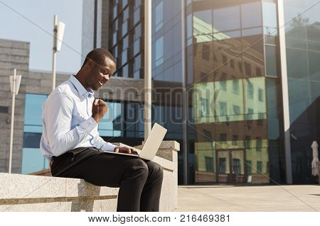 Happy black businessman working on laptop. African-american insuranse agent checking email on computer, talking on mobile, sitting on the bench outdoors near modern office center