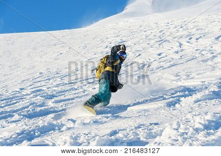 A freeboard snowboarder in a ski mask and a backpack skates over the snow-covered slope leaving behind a snow powder against the blue sky