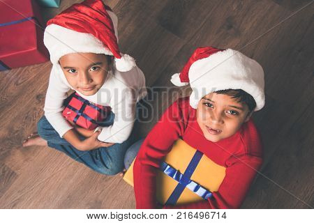 Indian kids celebrating christmas - cute little Indian kids playing, laughing and having fun with gift boxes on christmas, sitting on floor or sofa with christmas tree in the background