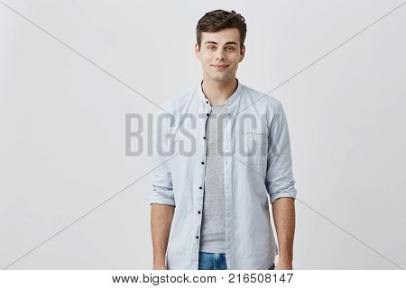 Cheerful pleased caucasian guy with blue eyes and dark hair dressed in elegant blue shirt smiling in good mood while posing in studio against gray background. Facial expressions concept