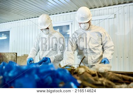 Portrait of two workers  wearing biohazard suits working at waste processing plant sorting recyclable materials on conveyor belt