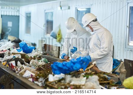Portrait of two workers  wearing biohazard suits and hardhats working at waste processing plant sorting recyclable materials on conveyor belt
