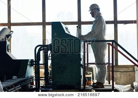 Side view full length of worker wearing biohazard suit operating massive machine in factory workshop standing against window