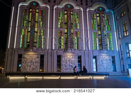 BERLIN GERMANY - AUGUST 26 2017; Two young women in dark of niight sit on bench below beautifully designed and lit Freidrichestrasse Theater