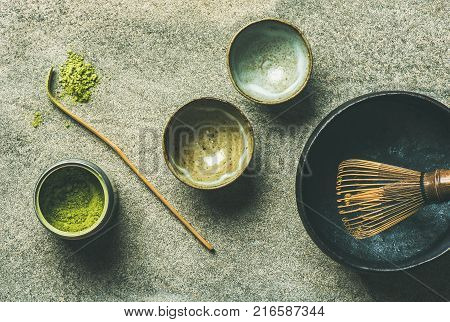 Flat-lay of Japanese tools for brewing matcha green tea. Matcha powder in tin can, Chashaku spoon, Chasen bamboo whisk, Chawan bowl and cups over grey background, top view