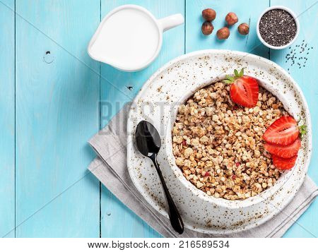 Top view of bowl with muesli, fresh strowberry, nuts, chia seeds and milk in milk jug on blue wooden background. Copy space.