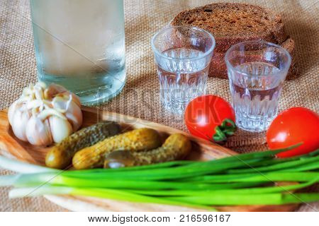 Vodka shot glasses and homemade lard with condiments. Fresh rye bread and pickled cucumbers. Garlic and vegetables. Rustic background. Traditional dishes of Russian cuisine. Close-up. Selective focus.