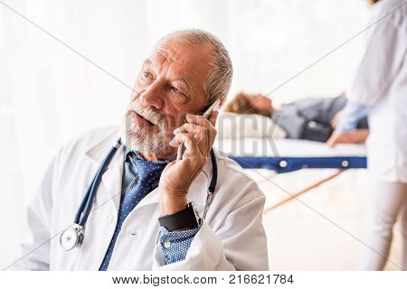 Senior doctor with smartphone in his office, making a phone call. A nurse and a lying patient in the background.