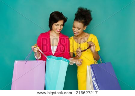 Happy smiling multiethnic girlfriends with colorful shopping bags. Two excited shopaholics boasting with purchases at turquoise sudio background with copy space