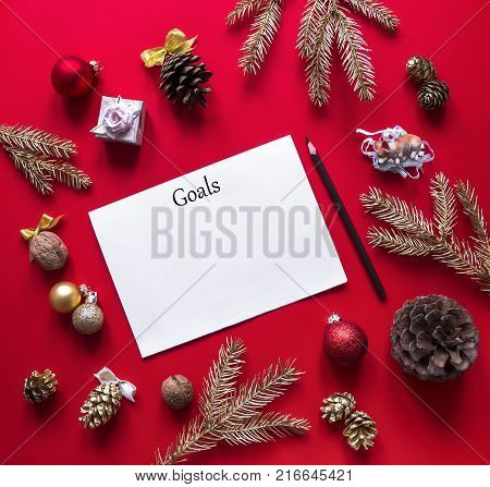 on a red raspberry background, New Year and Christmas decorations and spruce gold branches are arranged in a circle, and in the center lies a white sheet and a pencil goals