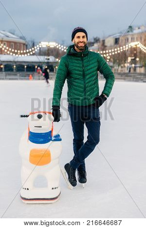 Outdoor Shot Of Glad Bearded Male Wears Green Jacket And Hat, Stands Near Skate Aid As Trying To Obt