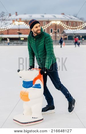 Outdoor Shot Of Unexperienced Male Skater Uses Skate Aid For Go Skating On Ice, Being In Good Mood,