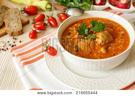 Traditional Georgian soup Kharcho with meat and rice in a white bowl next to bread, tomatoes, peppers, herbs on a white orange tablecloth. Horizontal. Close-up.