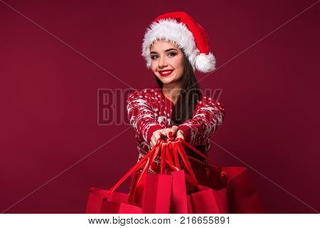 Young pretty woman or girl with long beautiful hair in Santa hat and New Year's dress holding red package on red studio background. Christmas sale. Emotional brunette. Copy space