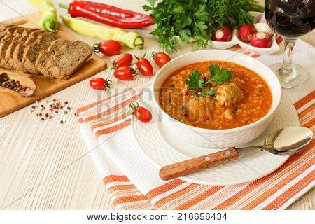 Traditional Georgian soup Kharcho with meat and rice in a white bowl next to bread, tomatoes, peppers, radish, herbs, a glass of red wine on a white orange tablecloth. Horizontal.