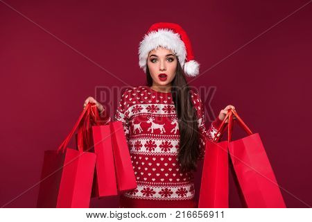 Young pretty woman or girl with long beautiful hair in Santa hat and New Year's dress holding red package on red studio background. Christmas sale. Emotional brunette. Copy space