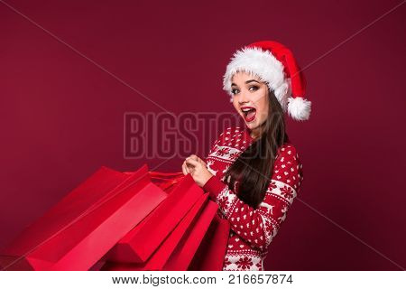 Young pretty woman or girl with long beautiful hair in Santa hat and New Year's dress holding red package on red studio background. Christmas sale. Emotional brunette. Copy space