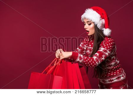 Young pretty woman or girl with long beautiful hair in Santa hat and New Year's dress holding red package on red studio background. Christmas sale. Emotional brunette. Copy space