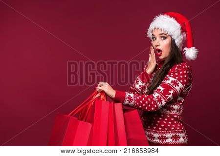 Young pretty woman or girl with long beautiful hair in Santa hat and New Year's dress holding red package on red studio background. Christmas sale. Emotional brunette. Copy space