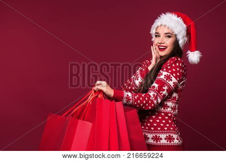 Young pretty woman or girl with long beautiful hair in Santa hat and New Year's dress holding red package on red studio background. Christmas sale. Emotional brunette. Copy space