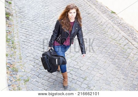 Focused young woman walking down street and carrying gyg bag with guitar. Female guitar player going to work in bar or club. Creative work concept.