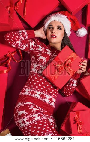 Funny young woman in a New Year's dress lying on a mountain of gifts. Christmas sale. Emotional brunette in Santa hat